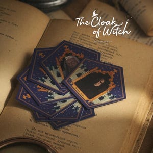 May include: A collection of pentagonal stickers featuring portraits of characters in ornate gold frames against a dark blue background with stars. The text The Cloak of Witch is printed in white script above the stickers on an open vintage book.