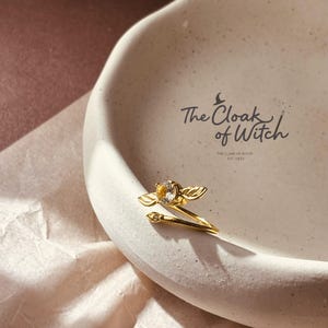 Könnte beinhalten: Ein goldfarbener Ring mit einem klaren Edelstein und flügelartigen Akzenten. Der Ring liegt auf einer weißen, strukturierten Keramikschale. Auf der Schale steht "The Cloak of Witch".
