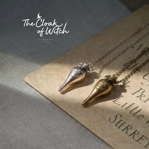 Könnte beinhalten: Zwei Halsketten mit Anhängern in Form von Tränkenflaschen, eine silberfarbene und eine goldfarbene, auf Vintage-Papier. Die Ketten haben feine Ketten und sind mit "The Cloak of Witch" und "EST 2022" beschriftet. Der Hintergrund ist in sanften Farben gehalten.