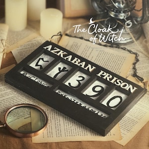 May include: A rectangular black wooden sign featuring the white text AZKABAN PRISON above a row of four square cutouts containing individual white tiles with black symbols and the number 390. The sign includes a small metal chain attached to the top.