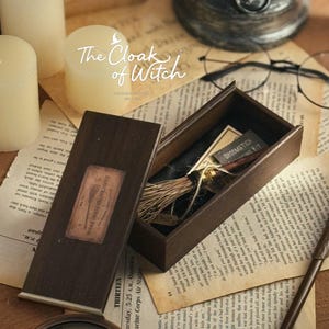 May include: A dark wooden rectangular box with a removable lid, containing a miniature broomstick with a brown handle and light-coloured bristles. The lid features a rectangular label with the text Handcrafted Flying Broom.