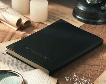 May include: A dark green book with gold-colored corner accents, lying on a tan surface with scattered aged paper. The book is surrounded by white candles, a magnifying glass, and a vintage lantern. The text "The Cloak of Witch" is visible.