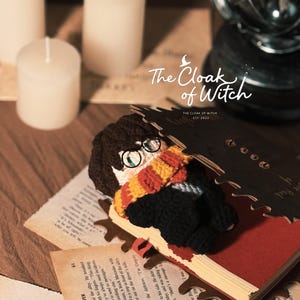 May include: A handmade crochet doll of a character with glasses and a scarf, lying on a book. The doll has brown hair and is wearing a black outfit. The image includes candles and text that reads "The Cloak of Witch".
