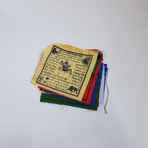 May include: A pile of prayer flags in various colours. The top flag is yellow with black text and illustrations. Other flags are red, green, blue, and white. A white string is attached.