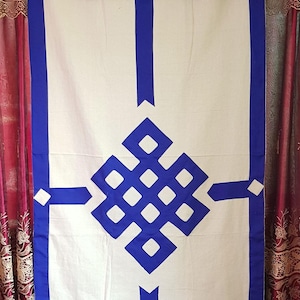 Op de afbeelding: Witte en blauwe decoratieve wandophanging met een geometrisch knoopontwerp. De bovenkant heeft een blauwe geplooide valance met gele, rode en blauwe accenten. De randen en het centrale ontwerp zijn blauw omlijnd.