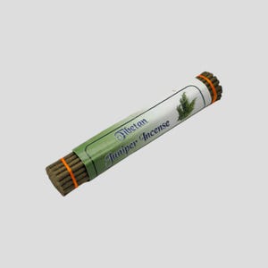 Könnte beinhalten: Ein Bündel tibetischer Wacholder-Räucherstäbchen, verpackt in einer grün-weißen Papierhülle. Die Hülle trägt den Text "Tibetan Juniper Incense" und eine Wacholderzweig-Illustration. Die Räucherstäbchen werden von einem orangefarbenen Gummiband zusammengehalten.