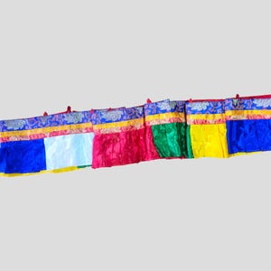 May include: A row of colorful prayer flags with a variety of colors including blue, white, red, green, and yellow. Each flag has a decorative patterned border in blue and gold.