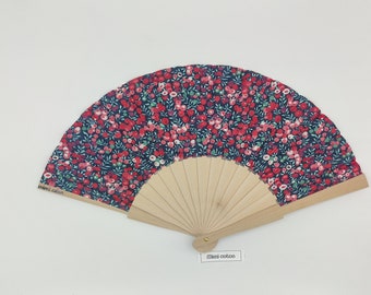 Op de afbeelding: Een handventilator met een bloemenpatroon. De stof van de ventilator heeft een donkerblauwe achtergrond met een herhalend patroon van rode en groene bloemen. Het houten frame van de ventilator is lichtbruin. De tekst "mimi coton" is op de ventilator gedrukt.
