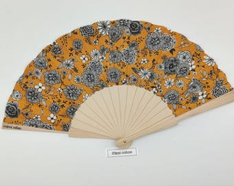 Op de afbeelding: Een handwaaier met een mosterdgele stoffen waaierblad voorzien van een zwart-wit bloemenpatroon. De waaier heeft lichtgekleurde houten baleinen en een metalen scharnierpunt. De tekst "mimi coton" staat op de linker balein en een label.