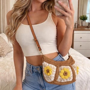 May include: A crochet shoulder bag with a brown strap and a floral design. The bag features a brown border with white and yellow sunflower-like flowers. The bag is worn over the shoulder.