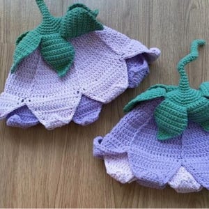 Op de afbeelding: Twee gehaakte hoeden in de vorm van klokvormige bloemen. Elke hoed heeft een lichaam met een kleurverloop van lichtpaars naar lavendel, een geschulpte rand en een donkergroene gehaakte steel- en bladstructuur bovenop de kroon.