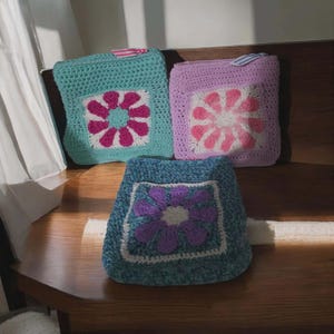 May include: Three handmade crochet pouches in shades of teal, lavender, and blue. Each pouch features a square floral design in contrasting colors: pink, white, and purple. The pouches are placed on a wooden surface, showcasing the handcrafted details.