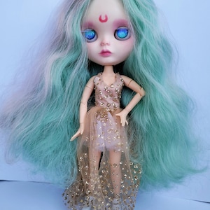 May include: A doll with long, wavy, teal and white hair, wearing a gold and pink dress with glitter. The doll has blue eyes and a red crescent moon painted on its forehead.