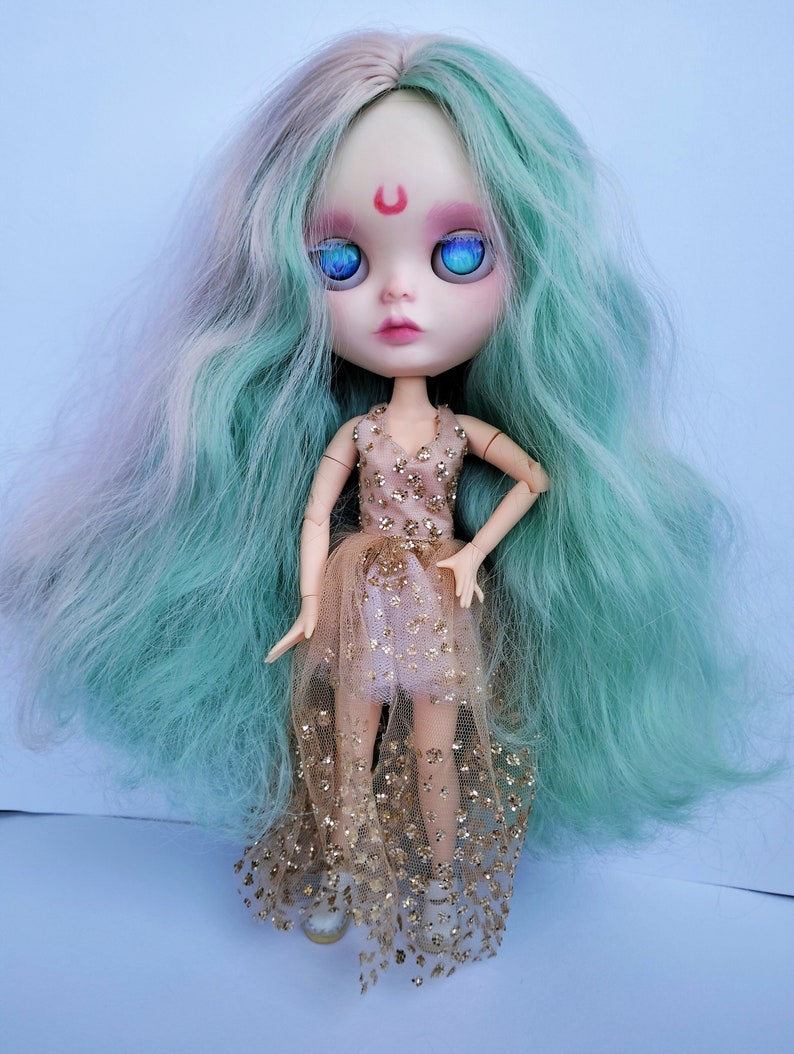 May include: A doll with long, wavy, teal and white hair, wearing a gold and pink dress with glitter. The doll has blue eyes and a red crescent moon painted on its forehead.