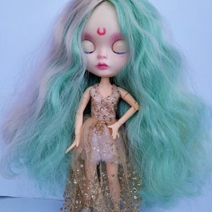 May include: A doll with long, wavy, teal and pink hair, wearing a gold glitter tulle dress and white shoes. The doll has closed eyes and a crescent moon painted on its forehead.