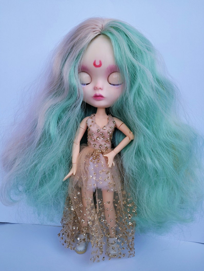 May include: A doll with long, wavy, teal and pink hair, wearing a gold glitter tulle dress and white shoes. The doll has closed eyes and a crescent moon painted on its forehead.