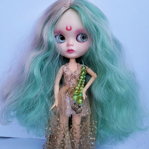 May include: A doll with long, wavy, teal and pink hair, wearing a gold and pink dress with a green beaded necklace and a heart charm.
