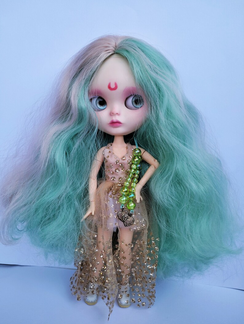 May include: A doll with long, wavy, teal and pink hair, wearing a gold and pink dress with a green beaded necklace and a heart charm.