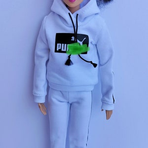 Può includere: Una bambola bianca che indossa una felpa con cappuccio bianca con bordi neri e la scritta "PUMA" sul davanti. La bambola indossa anche pantaloni della tuta bianchi con strisce nere sui lati e scarpe da ginnastica rosa.