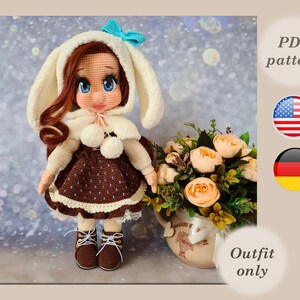 May include: Crochet doll pattern in PDF format, available in English and German. The doll is wearing a brown dress with white trim and a white hooded coat with bunny ears. The doll has brown hair and blue eyes.