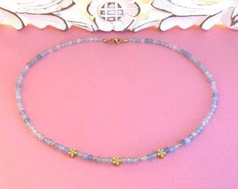 May include: A delicate blue and white beaded necklace with three gold flower charms. The necklace is on a pink background.