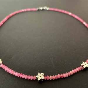 May include: A pink gemstone necklace with three silver flower charms. The necklace is made of small, faceted beads.
