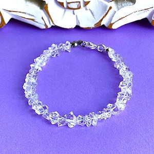 May include: A clear quartz crystal bracelet with a silver clasp, resting on a purple surface. The bracelet is composed of irregularly shaped, faceted quartz beads. The background includes a white decorative element.