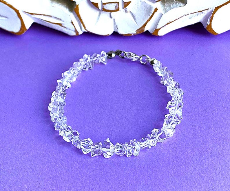 May include: A clear quartz crystal bracelet with a silver clasp, resting on a purple surface. The bracelet is composed of irregularly shaped, faceted quartz beads. The background includes a white decorative element.