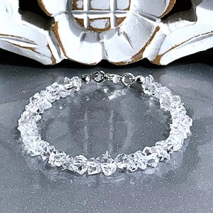 May include: A clear quartz crystal bracelet. The bracelet is made of small, irregularly shaped quartz crystals strung together with a silver-colored clasp. The crystals are translucent and reflect light, creating a sparkling effect.