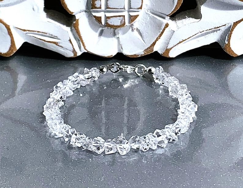 May include: A clear quartz crystal bracelet. The bracelet is made of small, irregularly shaped quartz crystals strung together with a silver-colored clasp. The crystals are translucent and reflect light, creating a sparkling effect.