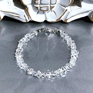 May include: A clear quartz crystal bracelet with a silver clasp. The bracelet is made of small, irregularly shaped quartz crystals strung together. The crystals are translucent and reflect light, creating a sparkling effect. The bracelet is displayed on a gray surface.