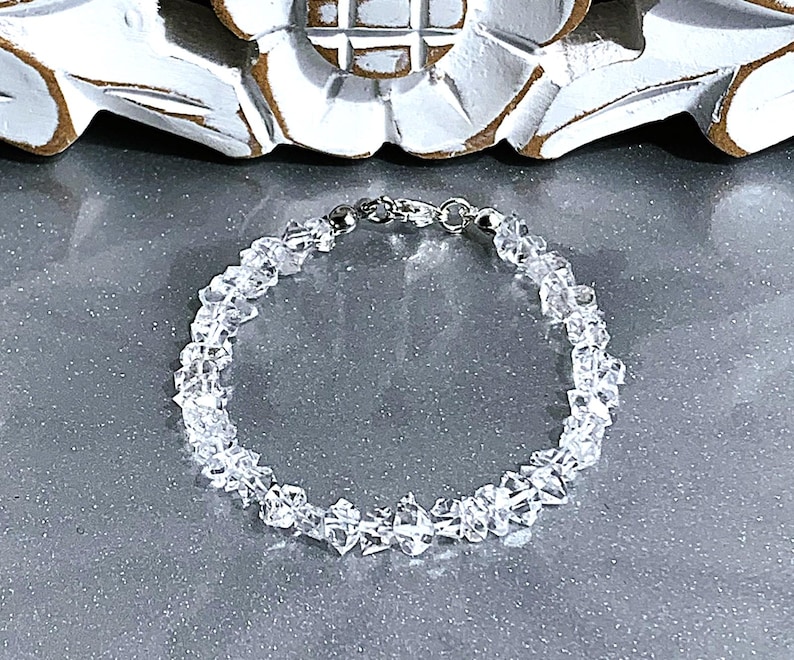 May include: A clear quartz crystal bracelet with a silver clasp. The bracelet is made of small, irregularly shaped quartz crystals strung together. The crystals are translucent and reflect light, creating a sparkling effect. The bracelet is displayed on a gray surface.