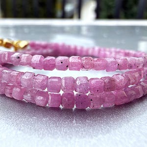 Op de afbeelding: Een armband met meerdere strengen, bestaande uit kleine, gefacetteerde, vierkant geslepen roze edelsteenkralen. De armband heeft een gepolijste goudkleurige metalen sluiting en is gerangschikt in drie afzonderlijke lussen, die de doorschijnende roze textuur tonen.