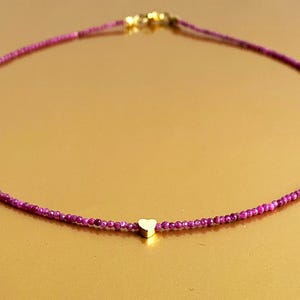 May include: A delicate gold heart charm hangs from a thin chain of tiny pink beads. The necklace is simple and elegant, perfect for everyday wear.