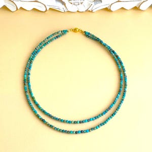 Op de afbeelding: Een turquoise kralenketting met twee strengen. De ketting heeft kleine, ronde kralen in blauwe en groene tinten, verbonden door een goudkleurige sluiting. De ketting wordt gepresenteerd op een lichtgele ondergrond.