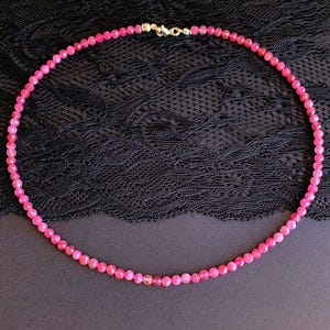 Può includere: Una delicata collana con piccole perline sfaccettate rosa acceso. Le perline sono infilate insieme per formare un cerchio completo, con una chiusura color oro. La collana è esposta su una superficie nera strutturata.