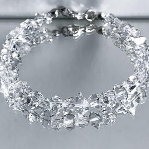 May include: A clear crystal bracelet with a silver clasp. The bracelet is made of many small, irregularly shaped crystals, giving it a sparkling appearance. The crystals are arranged in a circular pattern.
