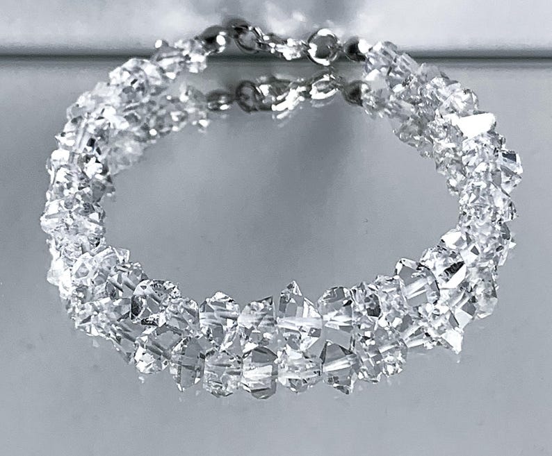 May include: A clear crystal bracelet with a silver clasp. The bracelet is made of many small, irregularly shaped crystals, giving it a sparkling appearance. The crystals are arranged in a circular pattern.