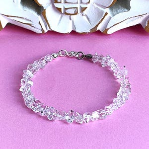 May include: Clear crystal bracelet with a silver clasp. The bracelet is made of irregularly shaped, clear crystals strung together. The background is a solid pink color, with a white and gold decorative element at the top.