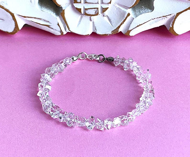 May include: Clear crystal bracelet with a silver clasp. The bracelet is made of irregularly shaped, clear crystals strung together. The background is a solid pink color, with a white and gold decorative element at the top.