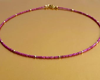 May include: A delicate necklace featuring a continuous strand of small, vibrant magenta-colored beads interspersed with tiny, polished gold-toned spherical beads. The necklace is secured with a small gold-toned metal clasp at the top center.