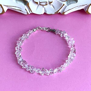 May include: A clear quartz crystal bracelet with a silver clasp, set against a pink background. The bracelet is composed of irregularly shaped, faceted quartz crystals. The crystals are strung together to form a circular bracelet.