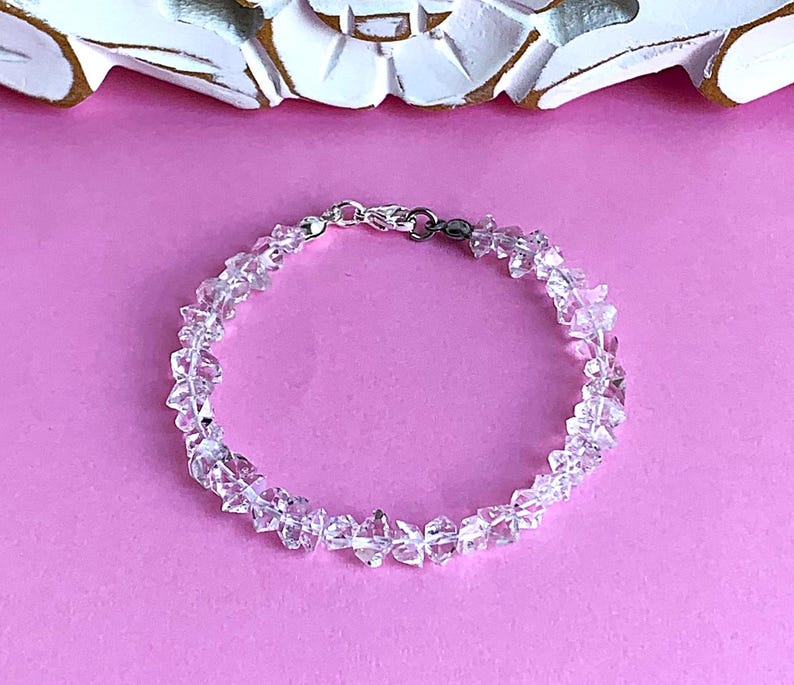 May include: A clear quartz crystal bracelet with a silver clasp, set against a pink background. The bracelet is composed of irregularly shaped, faceted quartz crystals. The crystals are strung together to form a circular bracelet.