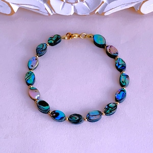 May include: Abalone shell bracelet with oval-shaped beads in shades of blue, green, and pink, strung together with gold-colored metal accents and a clasp. The bracelet is displayed on a light purple surface.