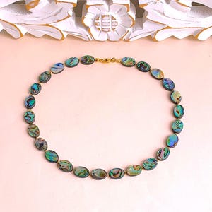 Könnte beinhalten: Halskette aus schillernden Abalone-Muschelkugeln in ovaler Form, die Farben von Blau, Grün und Gold zeigen. Die Perlen sind mit goldfarbenem Metall und einem Verschluss verbunden. Die Halskette wird auf einer hellrosa Oberfläche gezeigt.