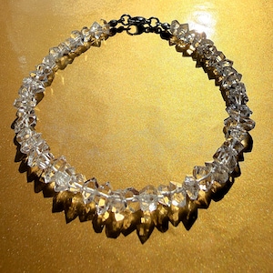 May include: A necklace composed of numerous clear, faceted, raw-cut crystal beads strung together in a circular arrangement. The piece features a dark metallic lobster claw clasp at the top, securing the strand of translucent, irregularly shaped stones.