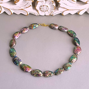 Puede incluir: Collar de concha de abulón iridiscente con cuentas ovaladas en tonos verdes, rosas y morados. Las cuentas están unidas con un cierre dorado. El collar se muestra sobre una superficie gris claro.