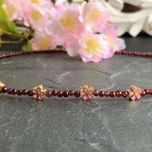 May include: A delicate red garnet necklace with small pink flower charms. The necklace is made of tiny red beads and features five gold flower charms with pink enamel.