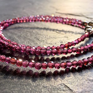 Könnte beinhalten: Eine zarte Halskette aus kleinen, facettierten, tiefroten Granatperlen. Die Kette verfügt an einem Ende über einen Metallverschluss, wobei die Perlen das Licht reflektieren und ihre durchscheinende, dunkelrote Farbe sowie ihre gleichmäßige Form zeigen.