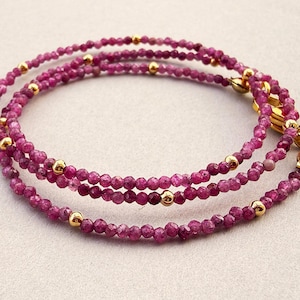Può includere: Una collana di perline a più fili con piccole perle di pietre preziose sfaccettate color rosa intenso, alternate a minuscole sfere di metallo lucido color oro. La collana è rifinita con una chiusura a moschettone dorata e una piccola targhetta rotonda.